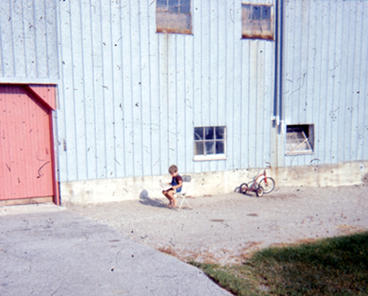 A little boy sits on a little chair in front of a metal barn reading in the sun.