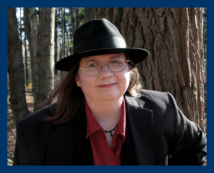 author photo wearing a black fedora & suit jacket over a red blouse in the autumn woods
