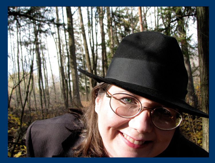author photo wearing a black fedora & suit jacket over a red blouse in the autumn woods